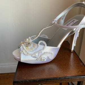 May include: A white satin T-strap high heel shoe featuring an open toe, a slim ankle strap, and a stiletto heel. The front of the shoe is decorated with intricate white embroidery, small white pearls, and a cluster of clear crystals.