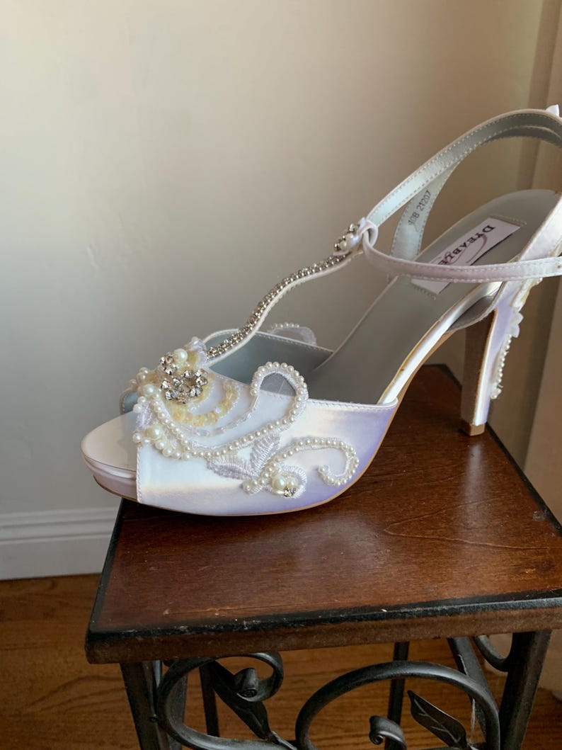 May include: A white satin T-strap high heel shoe featuring an open toe, a slim ankle strap, and a stiletto heel. The front of the shoe is decorated with intricate white embroidery, small white pearls, and a cluster of clear crystals.