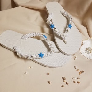 May include: White flip flops with a white crocheted strap decorated with blue starfish and white pearls. The flip flops are on a beige surface with small seashells scattered around.