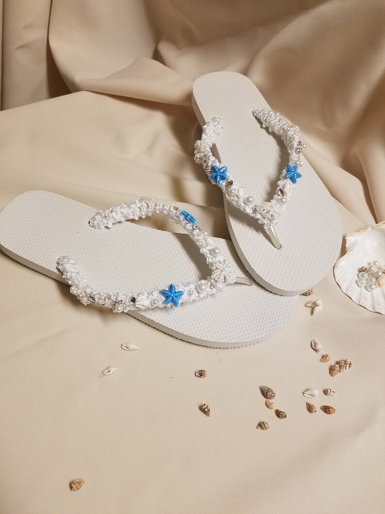 May include: White flip flops with a white crocheted strap decorated with blue starfish and white pearls. The flip flops are on a beige surface with small seashells scattered around.