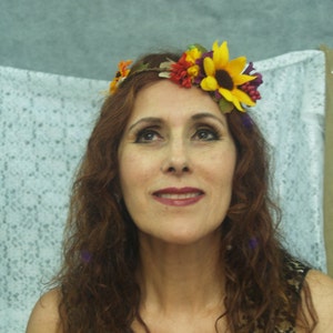 Op de afbeelding: Een vrouw met lang, krullend bruin haar draagt een bloemenkrans met gele, oranje en rode bloemen. De krans is gemaakt van bladeren en heeft een bruin lint.