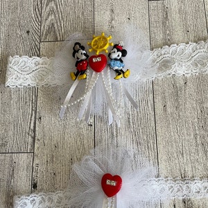 May include: Two white lace garters with red hearts and a white tulle bow. One garter features a Mickey Mouse and Minnie Mouse charm with a yellow steering wheel.