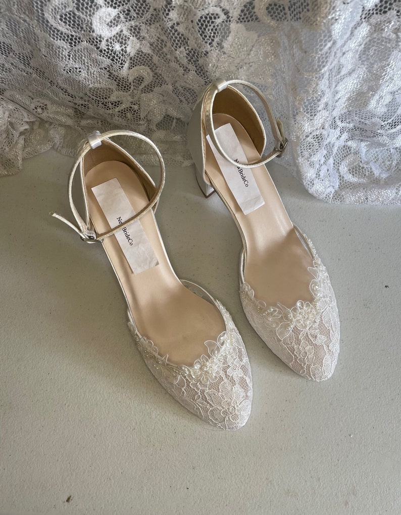 May include: A pair of ivory-colored bridal shoes with ankle straps and low heels. The toe of each shoe is adorned with delicate lace and small beads. The shoes have a label that reads "New BrideCo".