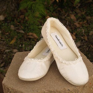 May include: Two pairs of white satin ballet flats with lace trim. The flats have a label that reads "NewBrideCo."