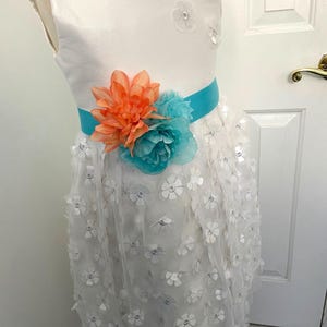 May include: White sleeveless dress with a turquoise and coral flower embellishment and a turquoise satin sash.