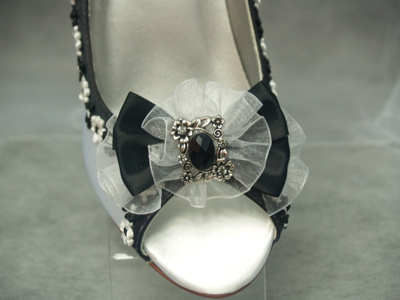 May include: White satin bridal shoes with a black satin bow and a silver and black jeweled embellishment.