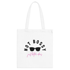 May include: White tote bag featuring the text "NOT BOSSY" in black, with black sunglasses and the pink script "just better ideas". The bag has two white handles, ideal for carrying items.