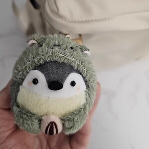 May include: A plush penguin toy wearing a green hooded outfit. The penguin has a gray body, white belly, and black eyes. The toy is round and fluffy, with a small, light-colored beak and a small, light-colored belly.