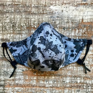 Könnte beinhalten: Eine schwarze und graue Gesichtsmaske mit einem Muster aus Schmetterlingen und Farbspritzern. Die Maske hat verstellbare Ohrschlaufen.
