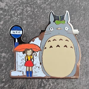 Puede incluir: Un pin de esmalte plateado que representa una escena de la película de animación Mi Vecino Totoro. El pin muestra a Totoro, una niña con un paraguas y una señal de parada de autobús con texto japonés. La niña lleva una camisa amarilla, una falda roja y botas azules.