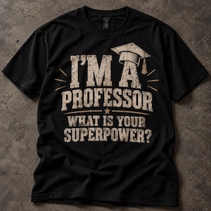 Op de afbeelding: Een zwart t-shirt met korte mouwen en een verweerde beige grafische opdruk met de tekst I'M A PROFESSOR WHAT IS YOUR SUPERPOWER? en een illustratie van een afstudeerhoed boven de tekst, gecentreerd op de voorzijde van het kledingstuk.