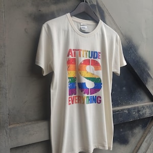 Puede incluir: Una camiseta de manga corta de color crema con un gran estampado gráfico de estilo desgastado. El texto dice "ATTITUDE IS EVERYTHING" con la letra "S" mostrada de forma prominente en un patrón de arcoíris en el centro del pecho.