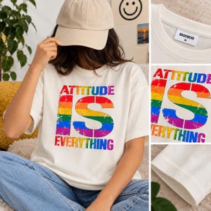 May include: A white short-sleeve t-shirt featuring the text "ATTITUDE IS EVERYTHING" in a distressed rainbow-coloured font. The shirt has a crew neckline and a "BASICWEAR M" label on the inside collar. The design is printed on the front centre.
