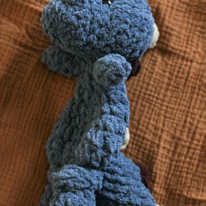 May include: A handmade blue crocheted dinosaur toy with a textured, plush appearance. The dinosaur has small arms and legs, a small head, and a dark brown patch on its back. The toy is lying on a brown fabric surface.