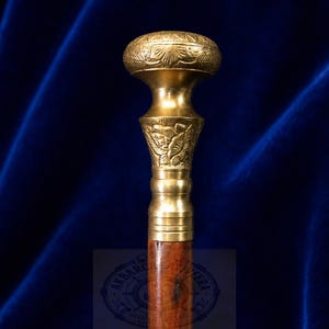 May include: A walking stick featuring a polished brass handle with an ornate, engraved mushroom-shaped top and a floral-patterned collar. The handle is attached to a smooth, dark brown wooden shaft, showcasing a clear, circular logo watermark.