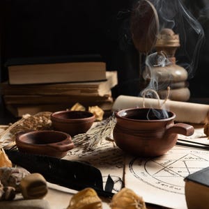 Puede incluir: Naturaleza muerta con una taza de cer&aacute;mica marr&oacute;n humeante, cuencos peque&ntilde;os y una pila de libros antiguos. La escena incluye elementos bot&aacute;nicos secos, una pluma negra y pergamino con s&iacute;mbolos ocultos, creando una atm&oacute;sfera m&iacute;stica.