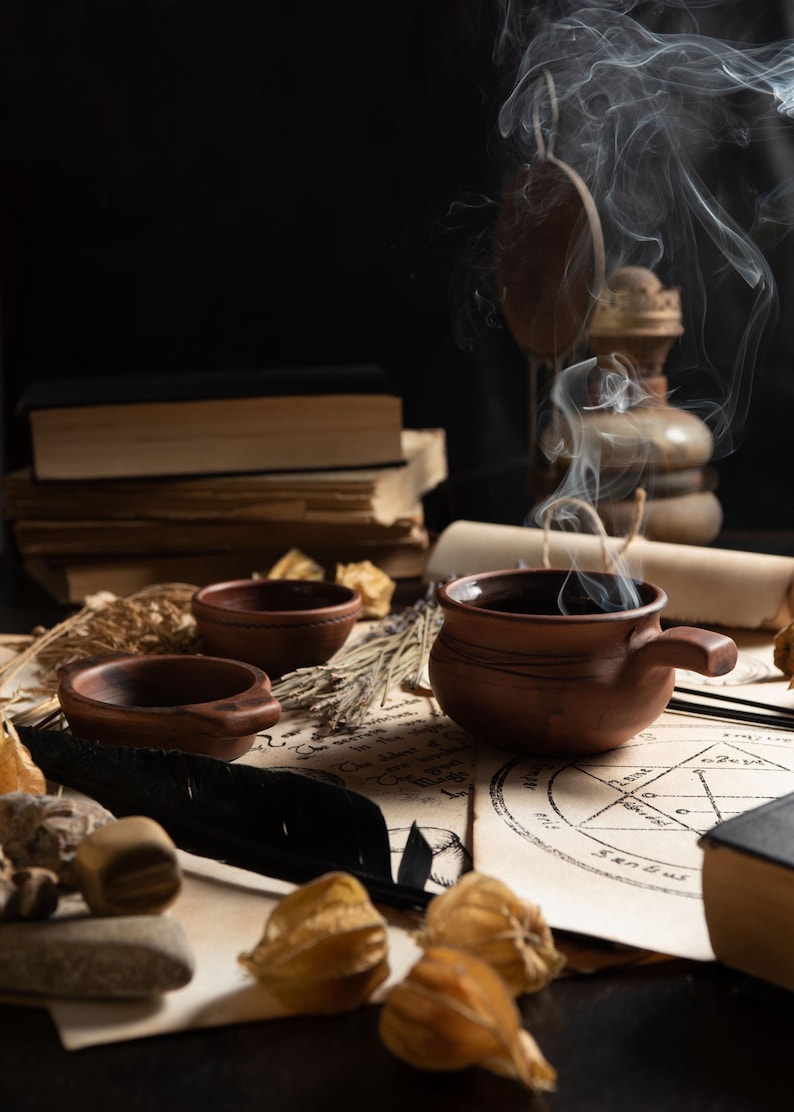 Puede incluir: Naturaleza muerta con una taza de cer&aacute;mica marr&oacute;n humeante, cuencos peque&ntilde;os y una pila de libros antiguos. La escena incluye elementos bot&aacute;nicos secos, una pluma negra y pergamino con s&iacute;mbolos ocultos, creando una atm&oacute;sfera m&iacute;stica.