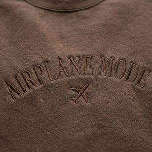 May include: A brown sweatshirt with the embroidered text "AIRPLANE MODE" in a curved arch above a small airplane graphic. The design is in a matching brown thread, creating a tone-on-tone effect.