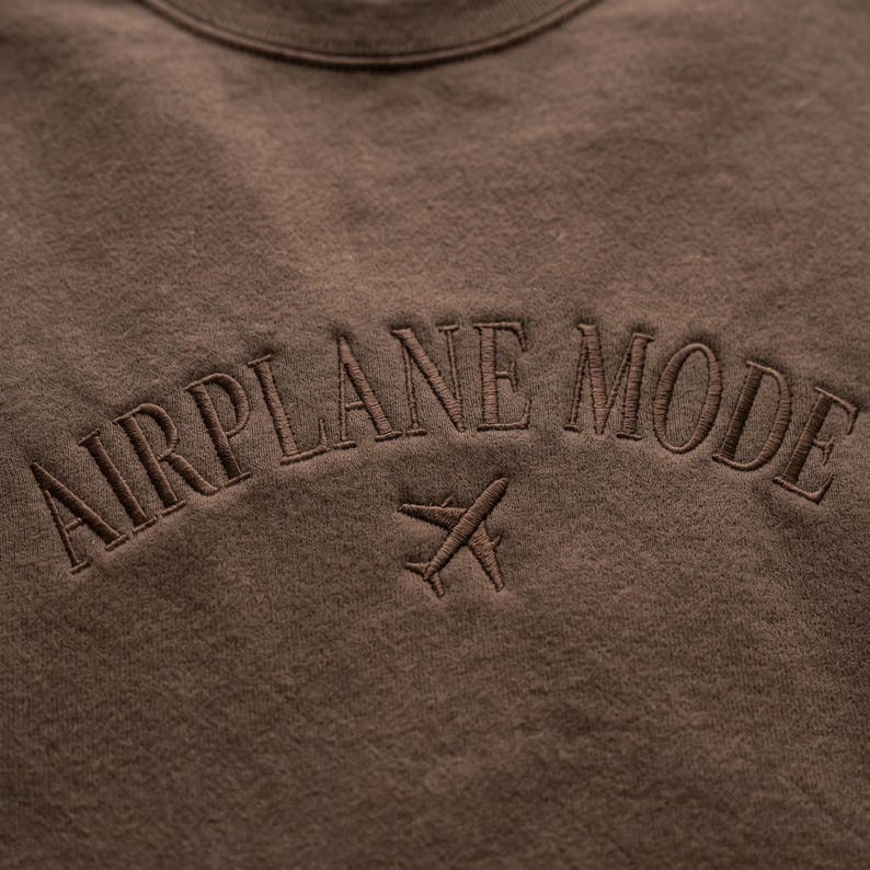 May include: A brown sweatshirt with the embroidered text "AIRPLANE MODE" in a curved arch above a small airplane graphic. The design is in a matching brown thread, creating a tone-on-tone effect.
