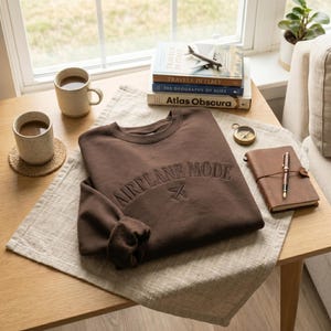 May include: A brown sweatshirt with the text "AIRPLANE MODE" embroidered across the chest, along with a small airplane icon. The sweatshirt is folded on a light-colored linen cloth, next to a stack of books and a leather-bound journal.