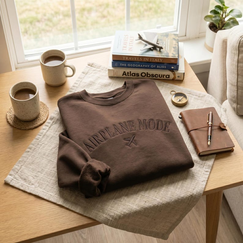 May include: A brown sweatshirt with the text "AIRPLANE MODE" embroidered across the chest, along with a small airplane icon. The sweatshirt is folded on a light-colored linen cloth, next to a stack of books and a leather-bound journal.