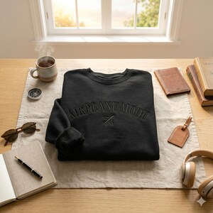 May include: A black sweatshirt with the words "AIRPLANE MODE" embroidered across the chest, along with a small airplane graphic. The sweatshirt is surrounded by travel accessories, including a passport cover, headphones, and a cup of tea.