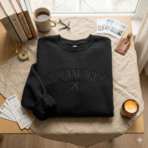 May include: Black crewneck sweatshirt with the text "AIRPLANE MODE" embroidered across the chest, along with a small airplane graphic. The sweatshirt is displayed on a wooden table with travel-themed accessories, including books, a compass, and a candle.