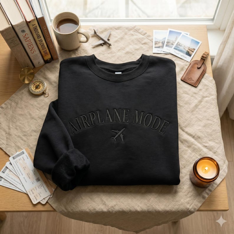May include: Black crewneck sweatshirt with the text "AIRPLANE MODE" embroidered across the chest, along with a small airplane graphic. The sweatshirt is displayed on a wooden table with travel-themed accessories, including books, a compass, and a candle.