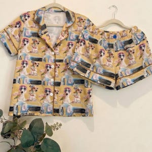 May include: A yellow pajama set featuring a repeating pattern of dogs wearing sunglasses. The set includes a button-down short-sleeved shirt and matching shorts. The pajamas are hanging on a white hanger against a light background.