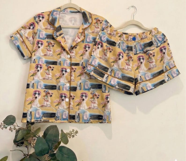 May include: A yellow pajama set featuring a repeating pattern of dogs wearing sunglasses. The set includes a button-down short-sleeved shirt and matching shorts. The pajamas are hanging on a white hanger against a light background.
