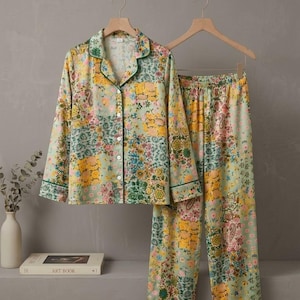 May include: A two-piece pajama set featuring a long-sleeve button-down shirt and matching pants. The fabric has a light green base with a colorful floral and paisley patchwork print, accented by dark green piping along the collar and cuffs.