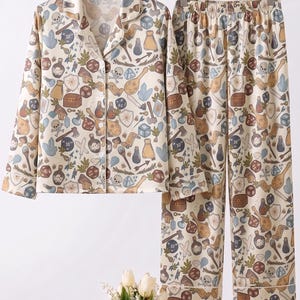 May include: Cream-colored pajama set with a repeating pattern of fantasy-themed icons. The long-sleeved top has a button-down front and notched collar. The pants are long and straight-legged. The pattern includes dice, potions, and shields.