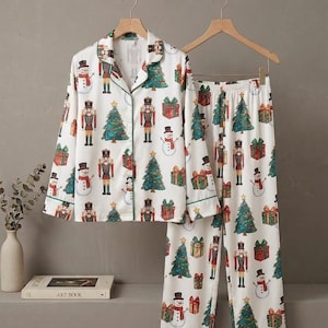 May include: A white pajama set featuring a long-sleeved button-down shirt with a notched collar and matching pants. The fabric is decorated with a repeating Christmas-themed print of nutcrackers, decorated pine trees, snowmen, and gift boxes.