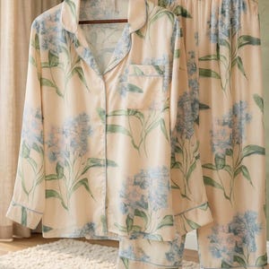 May include: A two-piece pajama set in a soft, cream color, adorned with a floral pattern in shades of blue and green. The long-sleeved top has a button-down front and a chest pocket. The matching pants complete the set.