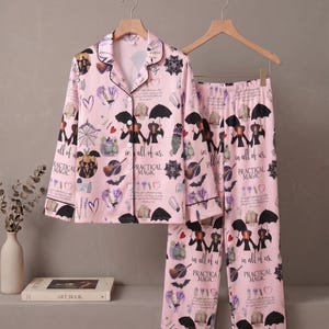 Puede incluir: Un conjunto de pijama rosa claro que incluye una camisa de manga larga con botones y un pantalón a juego. La tela presenta un patrón repetitivo de paraguas negros, telarañas, corazones y el texto "Practical Magic" y "in all of us."