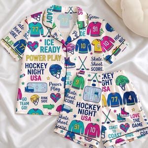 Puede incluir: Conjunto de pijama color crema con temática de hockey. La camiseta de manga corta y los pantalones cortos presentan camisetas de hockey, cascos, patines y texto como "Hockey Night USA" e "Ice Ready". El conjunto tiene ribetes negros y está decorado con varios colores.