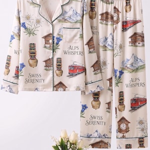 Può includere: Set pigiama color crema a tema Alpi svizzere. Il set comprende una camicia con bottoni e pantaloni, entrambi adornati con illustrazioni di montagne, chalet, campanacci e un treno rosso. La parte superiore e i pantaloni hanno finiture verdi e il testo "Swiss Serenity" e "Alps Whispers".