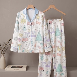 May include: White pajama set with a whimsical print of cakes, gifts, swans, and cars. The long-sleeved top has a light blue collar and cuffs. The matching pants have a relaxed fit. The set is hanging on a wooden hanger.