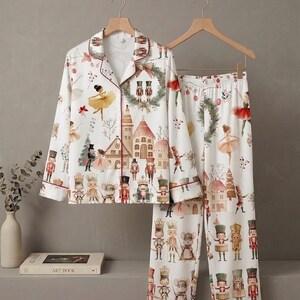 May include: A white long-sleeve pajama set featuring a festive print of nutcrackers, ballerinas, and holiday buildings. The button-down shirt has a notched collar with red piping, and the matching pants have an elastic waistband with a repeating pattern.