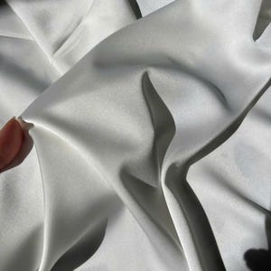 May include: Close-up of a white satin fabric with soft folds and shadows. The fabric has a smooth, lustrous appearance, reflecting light. The image highlights the texture and drape of the material, suggesting it's suitable for apparel or home decor.