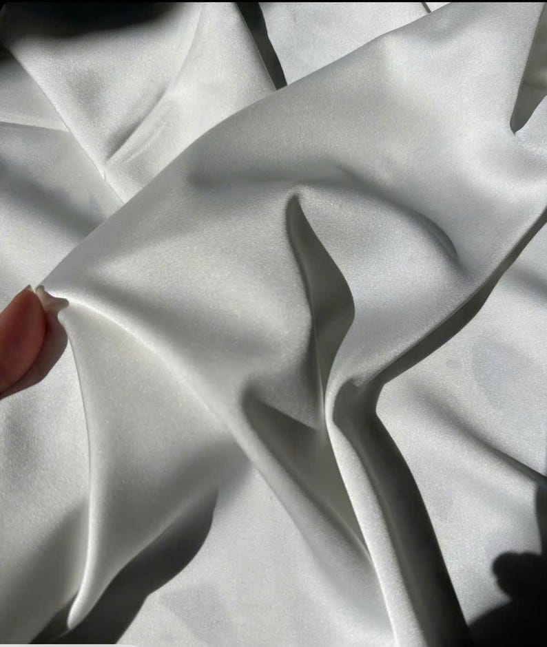 May include: Close-up of a white satin fabric with soft folds and shadows. The fabric has a smooth, lustrous appearance, reflecting light. The image highlights the texture and drape of the material, suggesting it's suitable for apparel or home decor.