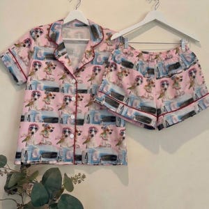May include: Pink pajama set featuring a repeating pattern of dogs wearing sunglasses and driving a blue car. The set includes a button-down short-sleeved shirt and matching shorts, both trimmed with a dark red piping.