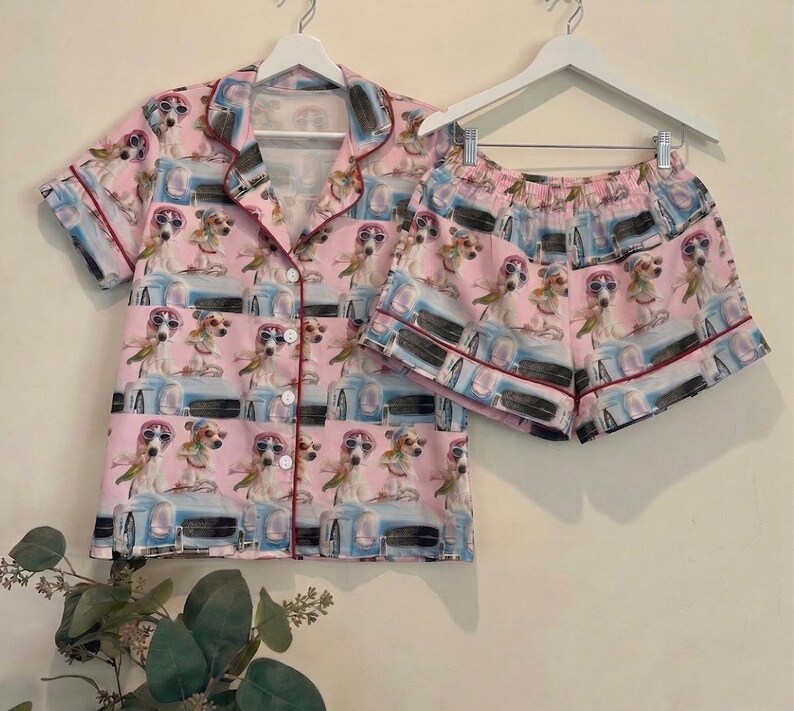 May include: Pink pajama set featuring a repeating pattern of dogs wearing sunglasses and driving a blue car. The set includes a button-down short-sleeved shirt and matching shorts, both trimmed with a dark red piping.