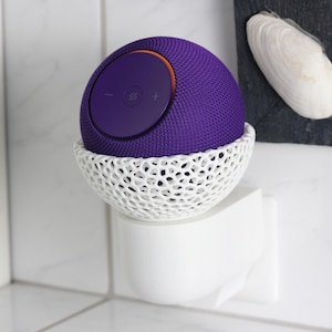 Puede incluir: Un altavoz esférico morado con un acento naranja se encuentra en un soporte calado blanco. El altavoz tiene botones de control de volumen. El soporte está montado en una toma de corriente blanca. Una concha marina está en el fondo.