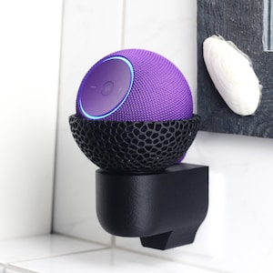 Puede incluir: Un altavoz inteligente morado en forma de esfera, alojado en un soporte negro con diseño. Está fijado a un soporte de pared negro. El altavoz tiene una parte superior circular con luz azul y un botón de encendido.