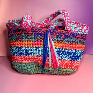 Könnte beinhalten: Eine farbenfrohe, handgefertigte Tragetasche mit Streifenmuster. Die Tasche weist eine Vielzahl leuchtender Farben auf, darunter Rosa, Blau, Orange und Grün. Sie hat geflochtene Griffe und dekorative Quasten. Die Tasche besteht aus einem weichen, gewebten Material.