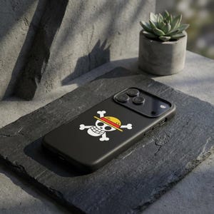May include: Black phone case featuring a white skull and crossbones design with a yellow and red straw hat. The case is made of a smooth material and is designed to fit a smartphone. The case is on a dark gray surface.
