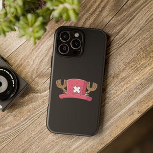 Könnte beinhalten: Schwarze Handyhülle mit einem rosa Hut, einem weißen Kreuz und braunen Geweihen. Das Telefon liegt auf einer Holzoberfläche, mit einer Kamera und Grünzeug im Hintergrund. Das Hutdesign ist im Cartoon-Stil gehalten.