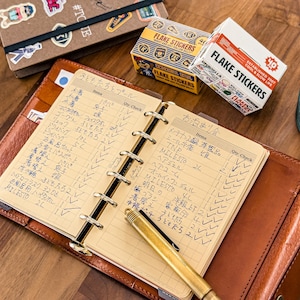 May include: A brown leather planner with a handwritten list, a gold pen, and boxes of flake stickers. The planner is open to a page with Japanese text and check boxes. A brown notebook with stickers is also visible.
