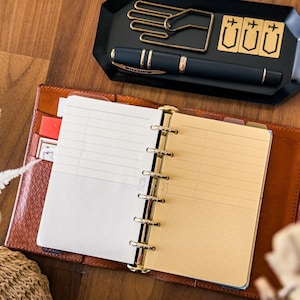 May include: Open brown leather planner with lined paper, gold-tone rings, and a pen in the side pocket. A black tray holds a pen, gold-tone paper clips, and decorative shapes. The planner is open on a wooden surface.